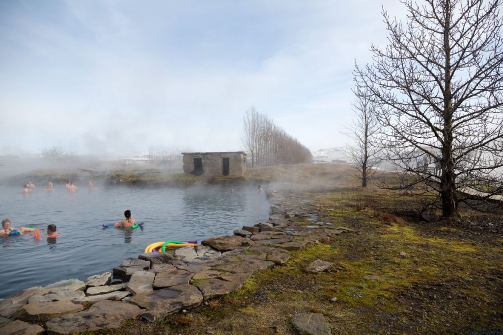 People soaking in the misty waters of the Secret Lagoon hot spring in Iceland, surrounded by natural stone paths and bare winter trees.