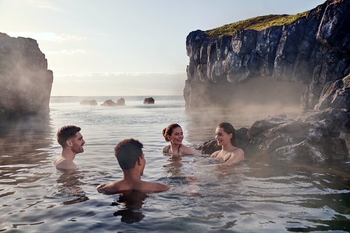 Four people relaxing in a natural geothermal hot spring surrounded by rocky cliffs and mist at Sky Lagoon in Iceland.