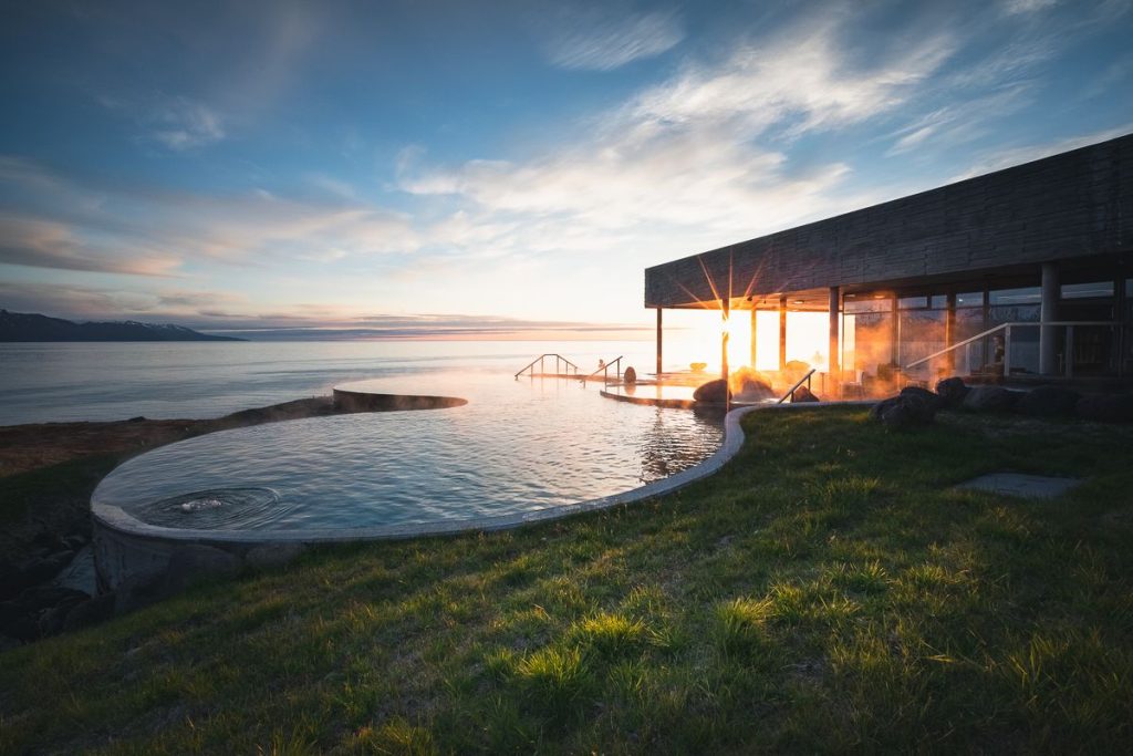 Infinity-style geothermal pool at sunset in Iceland, with steam rising and a modern spa building overlooking a calm fjord surrounded by mountains.