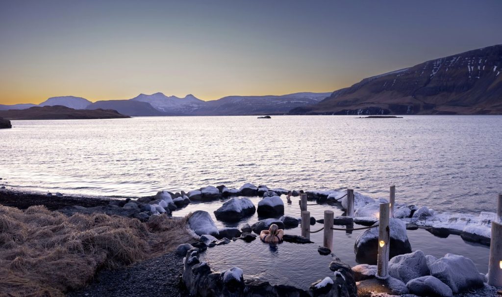 Person relaxing in a small natural hot spring overlooking the ocean at Hvammsvík Hot Springs, with snowy rocks and mountain views at sunset.