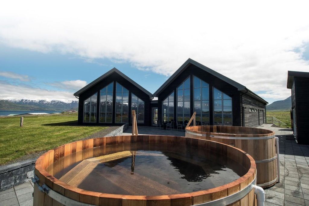 Wooden outdoor beer baths in front of a modern glass-fronted spa building in Iceland, with views of snow-capped mountains and a calm fjord in the background.