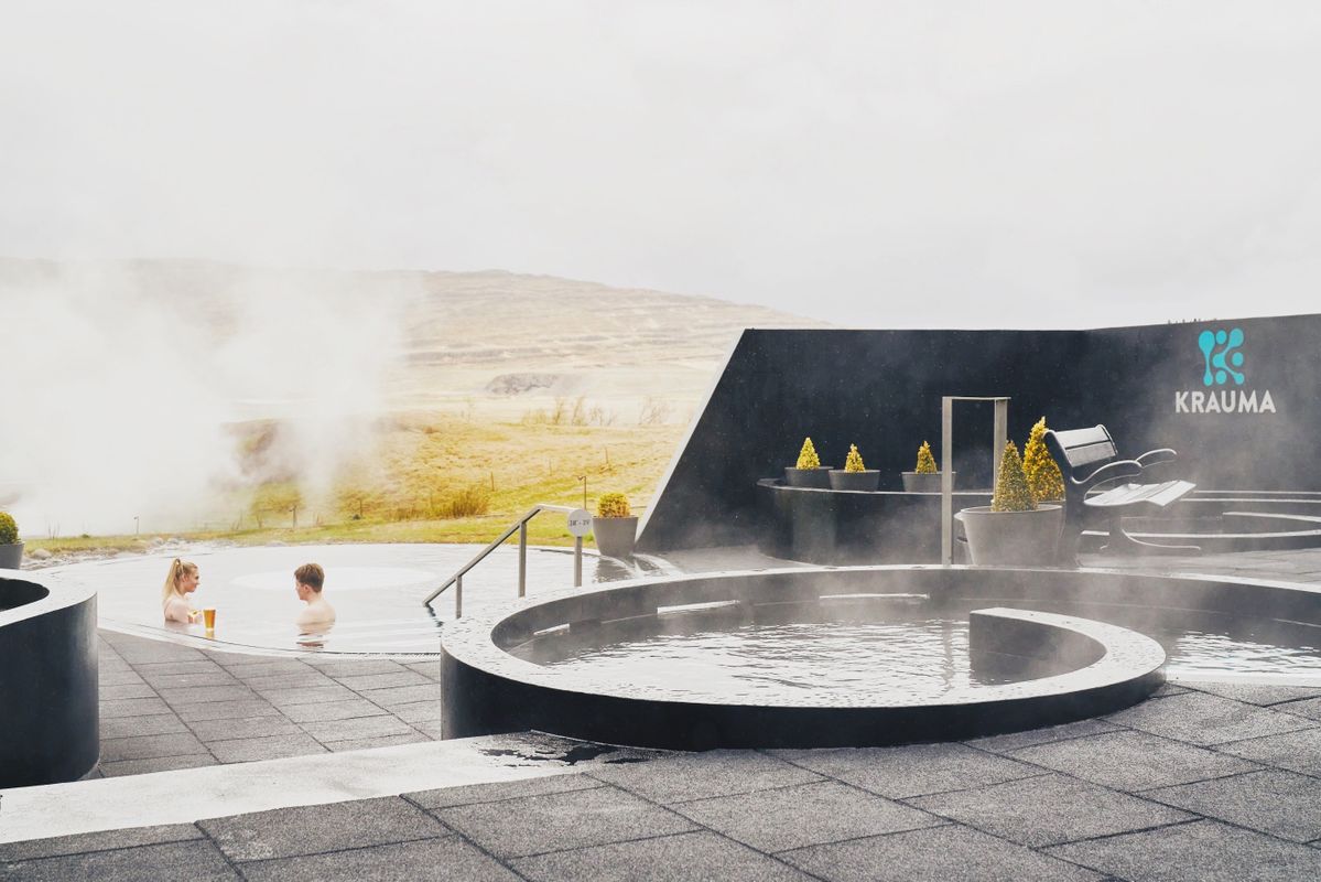 wo people relaxing in the Krauma geothermal baths with steam rising around them, surrounded by minimalist black architecture and natural Icelandic landscape in the background.