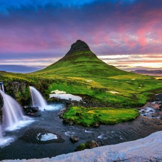Kirkjufell mountain at sunset with Kirkjufellsfoss waterfall in the foreground, surrounded by lush green fields and snow patches in Iceland.