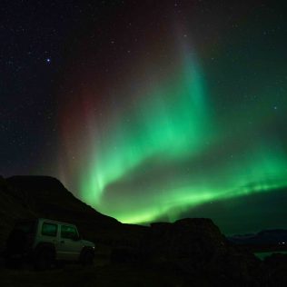 Northern Lights (Aurora Borealis) glowing green in the night sky over a rugged Icelandic landscape, with a 4x4 vehicle parked beneath the illuminated sky.