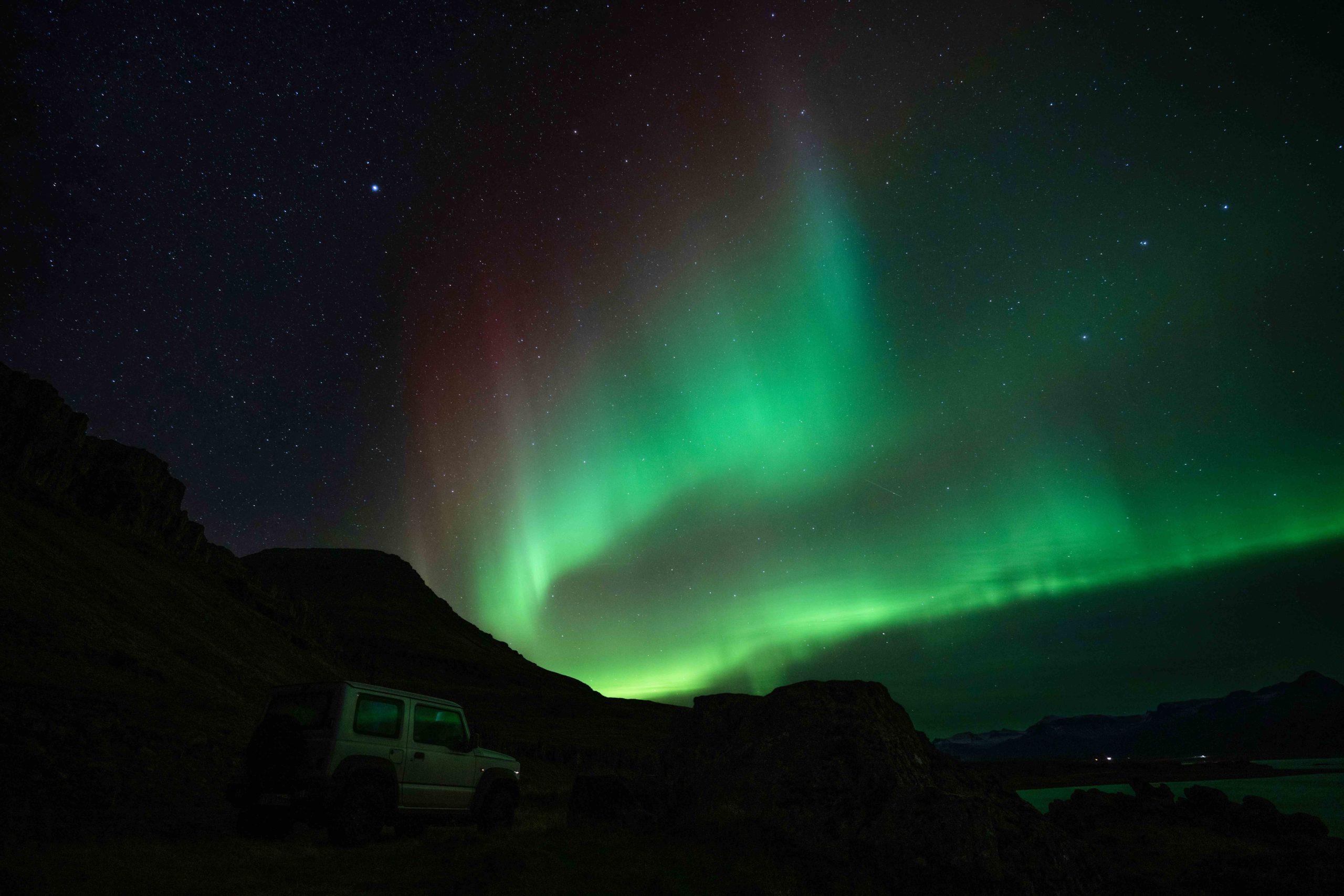 Northern Lights (Aurora Borealis) glowing green in the night sky over a rugged Icelandic landscape, with a 4x4 vehicle parked beneath the illuminated sky.