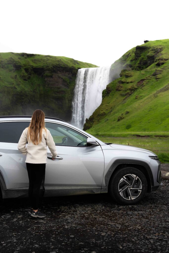 Woman standing beside silver rental car near Skógafoss waterfall, South Coast Iceland — Thrifty Car Rental Iceland self-drive trip.
